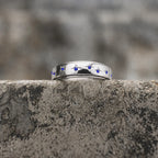 A polished silver ring featuring a row of small blue gemstone star motifs, resting on a textured gray stone ledge with a blurred natural background.