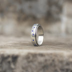 A polished silver band ring with small blue gemstone accents, standing upright on a textured stone surface with a blurred natural background.