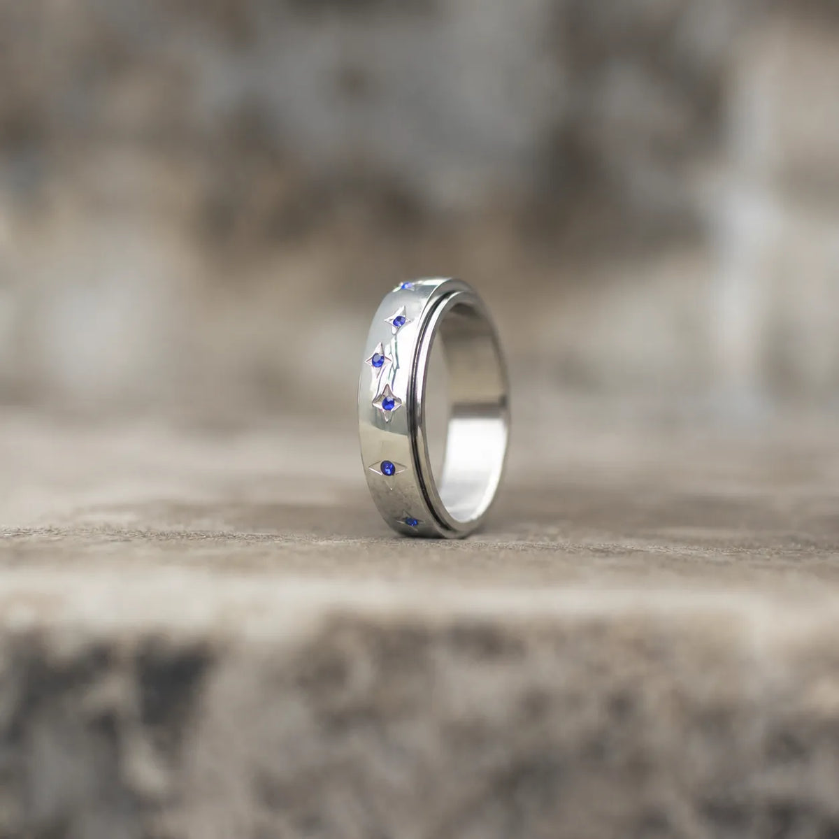A polished silver band ring with small blue gemstone accents, standing upright on a textured stone surface with a blurred natural background.