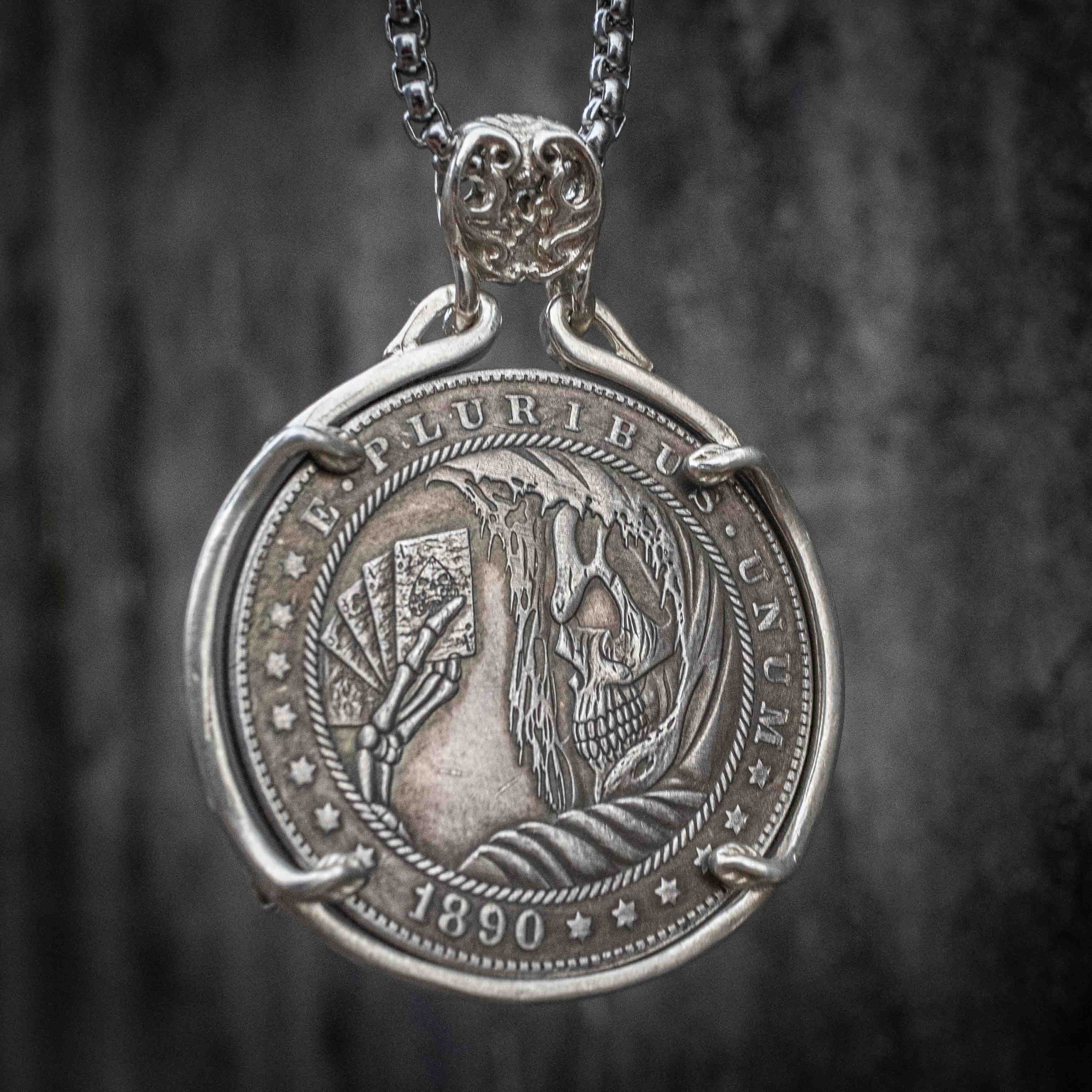 Coin pendant engraved with a skeleton holding playing cards amid flame and feather details, rope border, dated 1890, set in a four-prong bezel on a box chain Main image