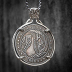 Coin pendant engraved with a skeleton holding playing cards amid flame and feather details, rope border, dated 1890, set in a four-prong bezel on a box chain