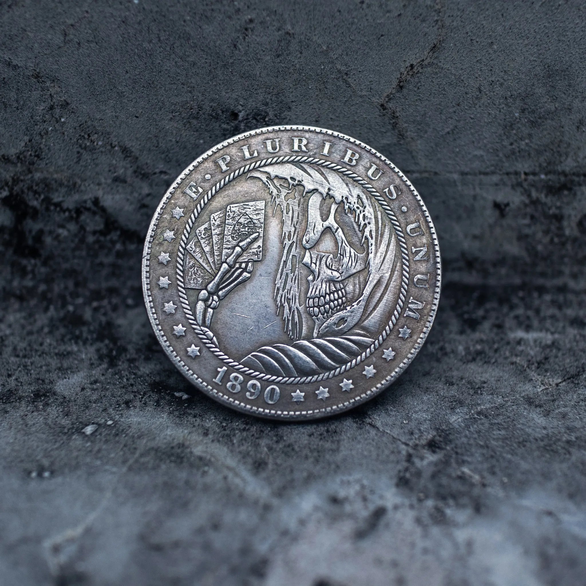 Close-up of a hobo nickel style coin engraved with a skeleton holding a fan of playing cards, flame and feather background, rope border, E Pluribus Unum, dated 1890