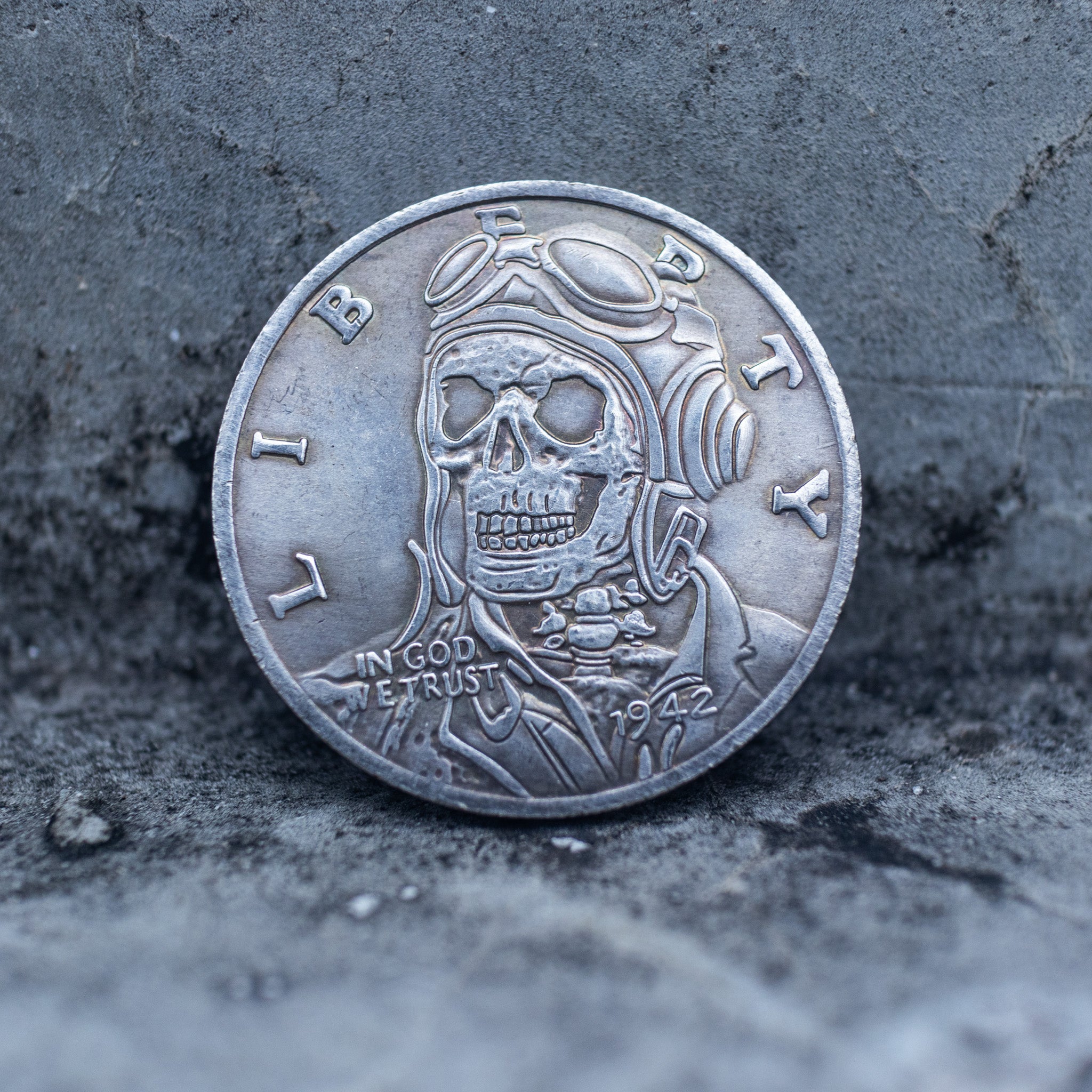 Close-up of a coin engraved with a skull in aviator goggles and flight jacket, Liberty and In God We Trust inscriptions, dated 1942, on dark stone