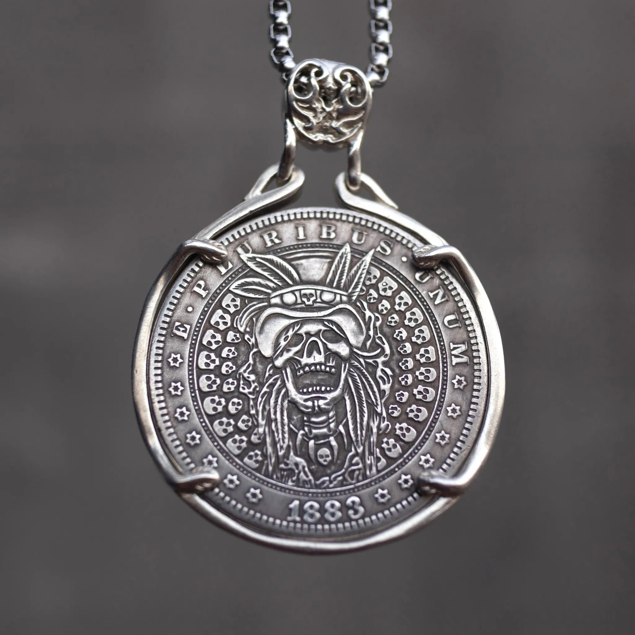 Coin pendant engraved with a skull wearing a Native American war bonnet headdress, surrounded by a border of small skulls, dated 1883, set in a four-prong bezel on a chain