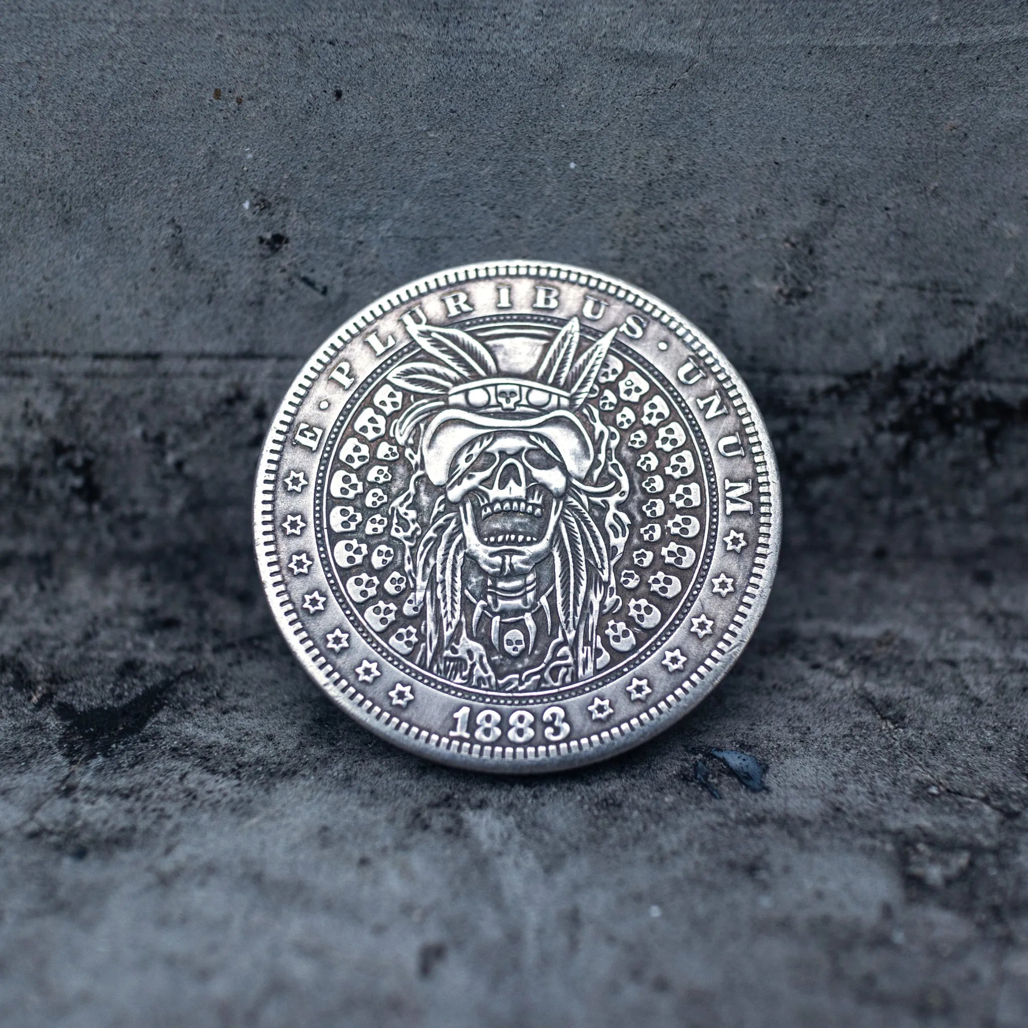 Close-up of a coin engraved with a skull in a feathered war bonnet, ring of small skulls as border decoration, E Pluribus Unum outer edge, dated 1883, on dark stone