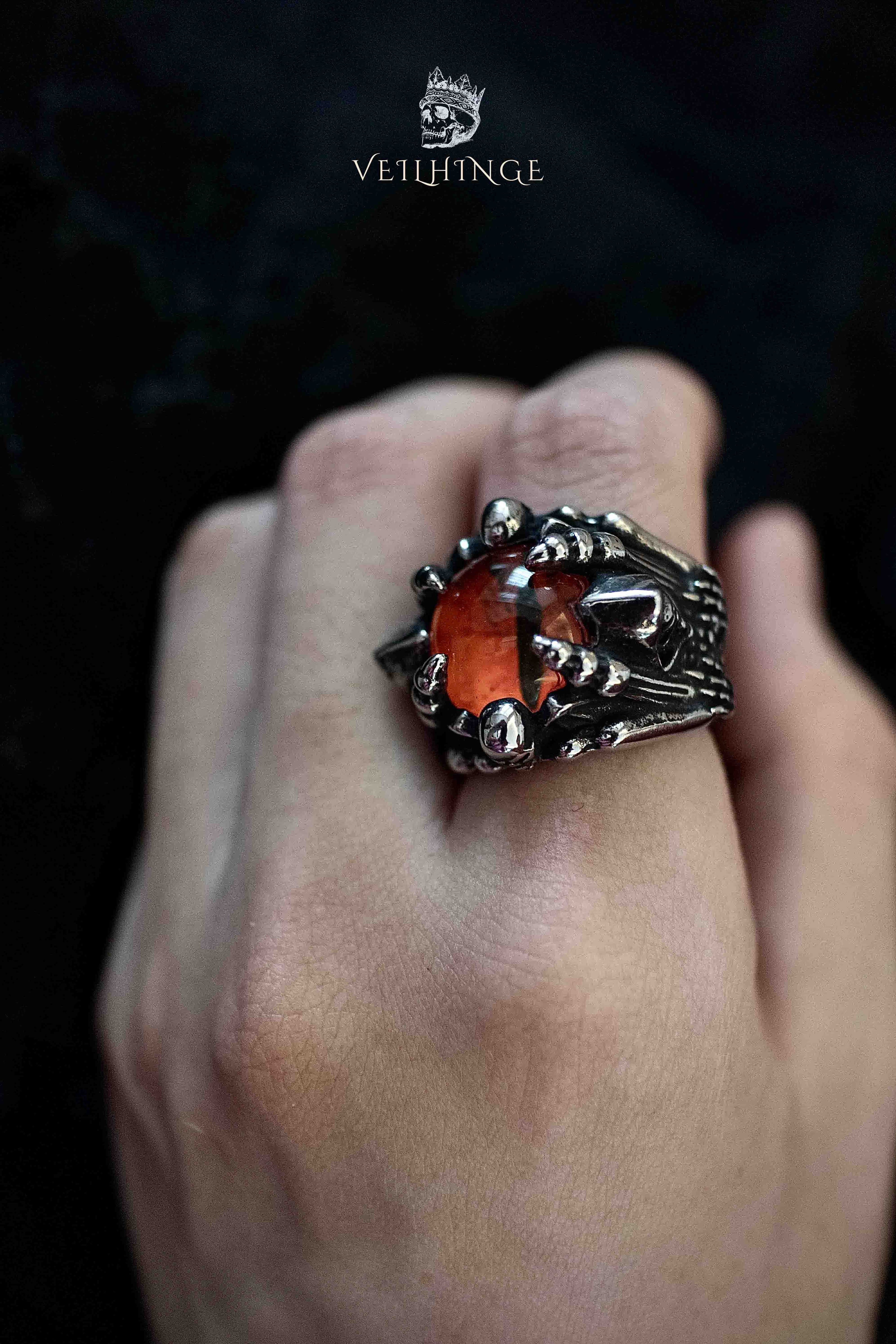 Side view of a dragon claw ring with an orange cushion-cut stone, hooded skeleton figures visible on the band shoulders, oxidized dark metal finish
