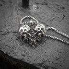 skull heart pendant laid flat on wet dark stone — twin skull design with worn-in oxidized finish