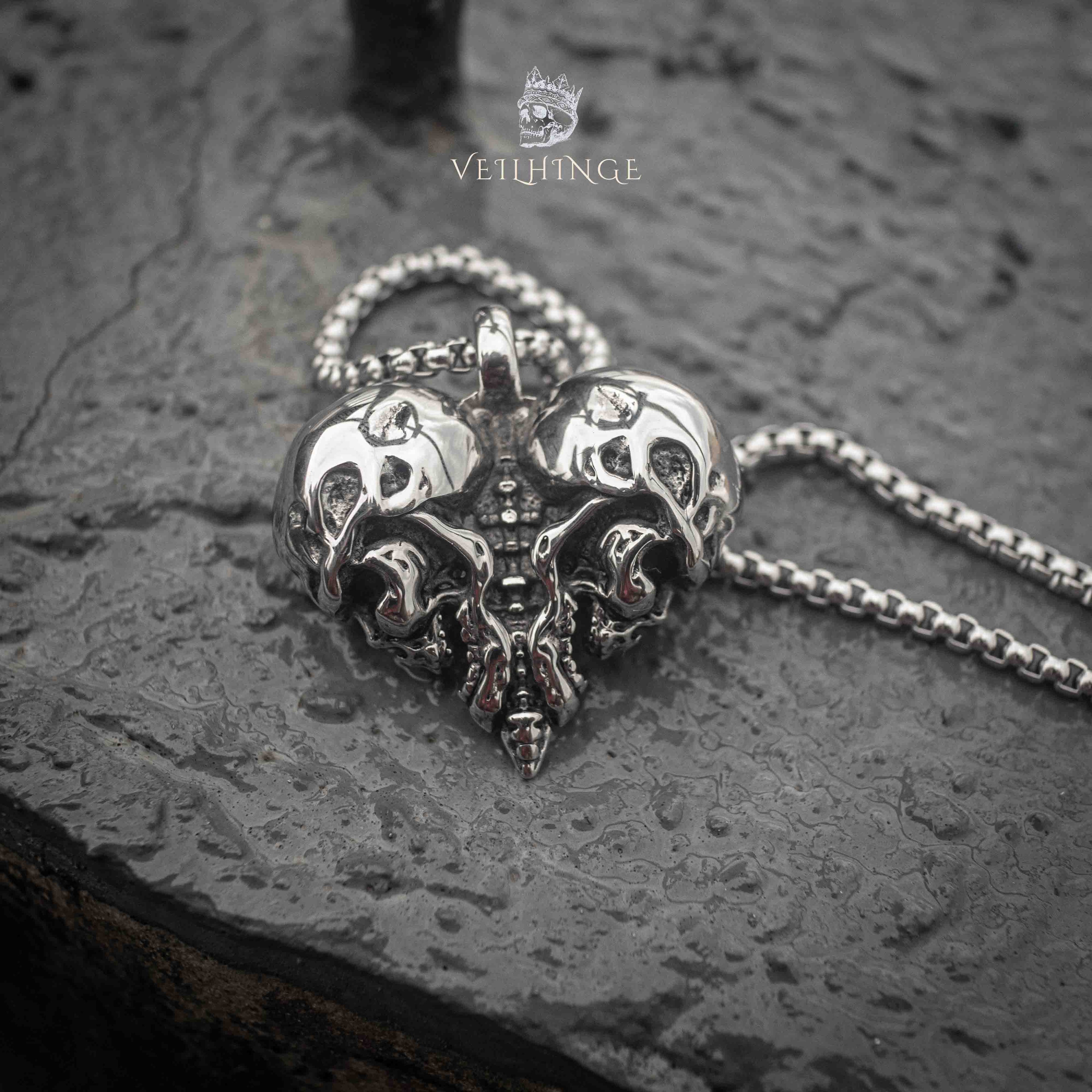 skull heart pendant laid flat on wet dark stone — twin skull design with worn-in oxidized finish