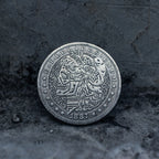 Close-up of a coin engraved with a skull-faced Lady Liberty, flowing curly hair, wheat and floral crown, striped shield below, E Pluribus Unum border, dated 1887