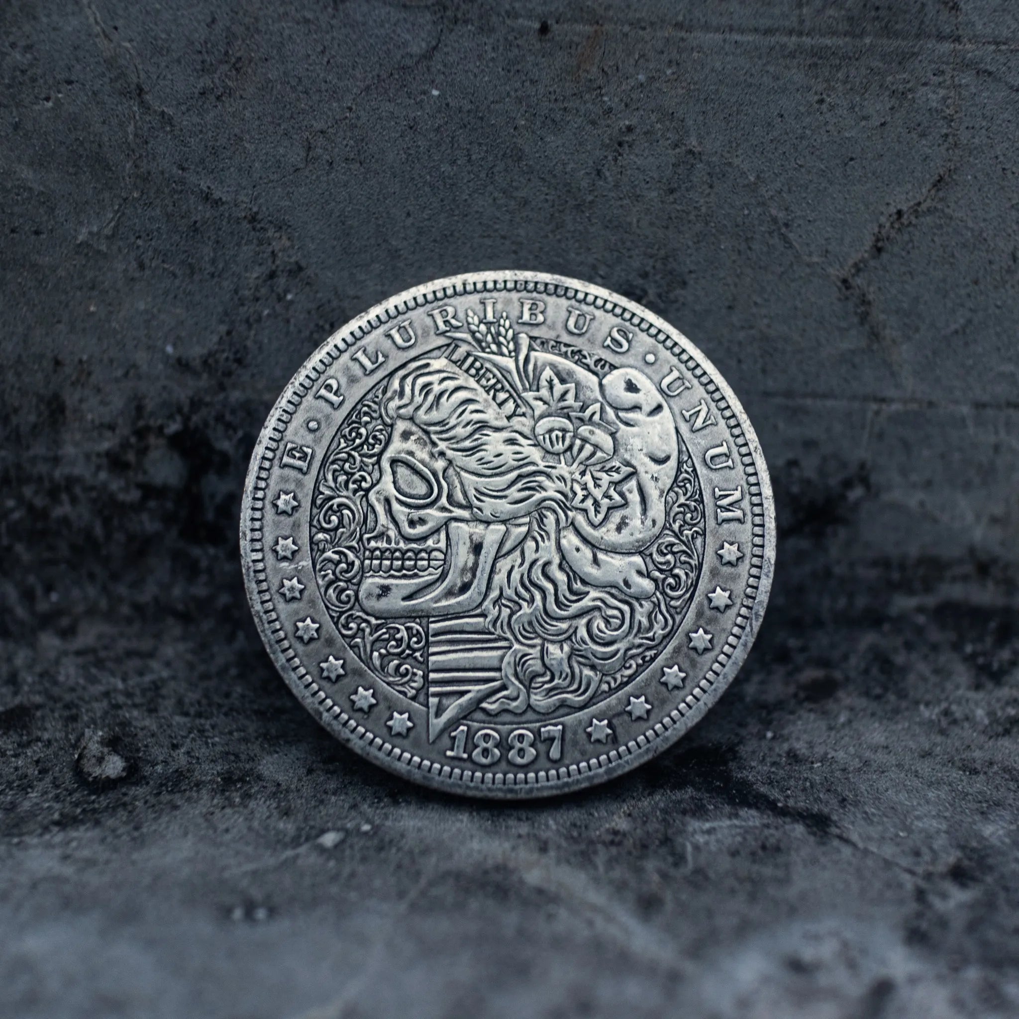 Close-up of a coin engraved with a skull-faced Lady Liberty, flowing curly hair, wheat and floral crown, striped shield below, E Pluribus Unum border, dated 1887