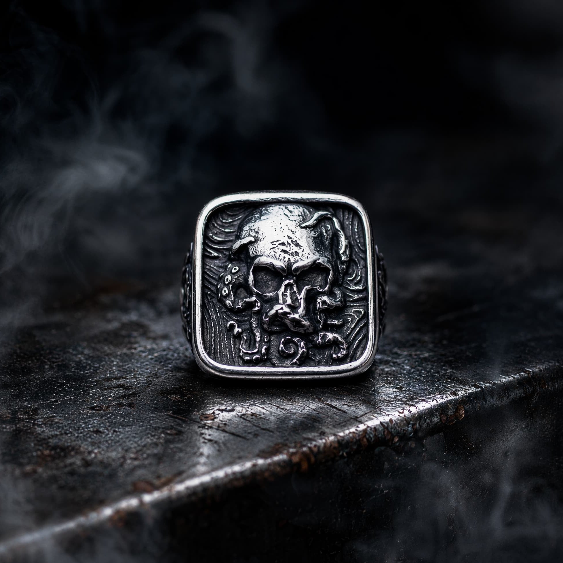 Square signet ring with skull centerpiece surrounded by octopus tentacles — oxidized silver relief on dark gothic signet face, wet black stone surface