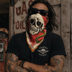 Tattooed man wearing a skull and snake printed bandana folded as a face covering, shot in front of a vintage gas station wall