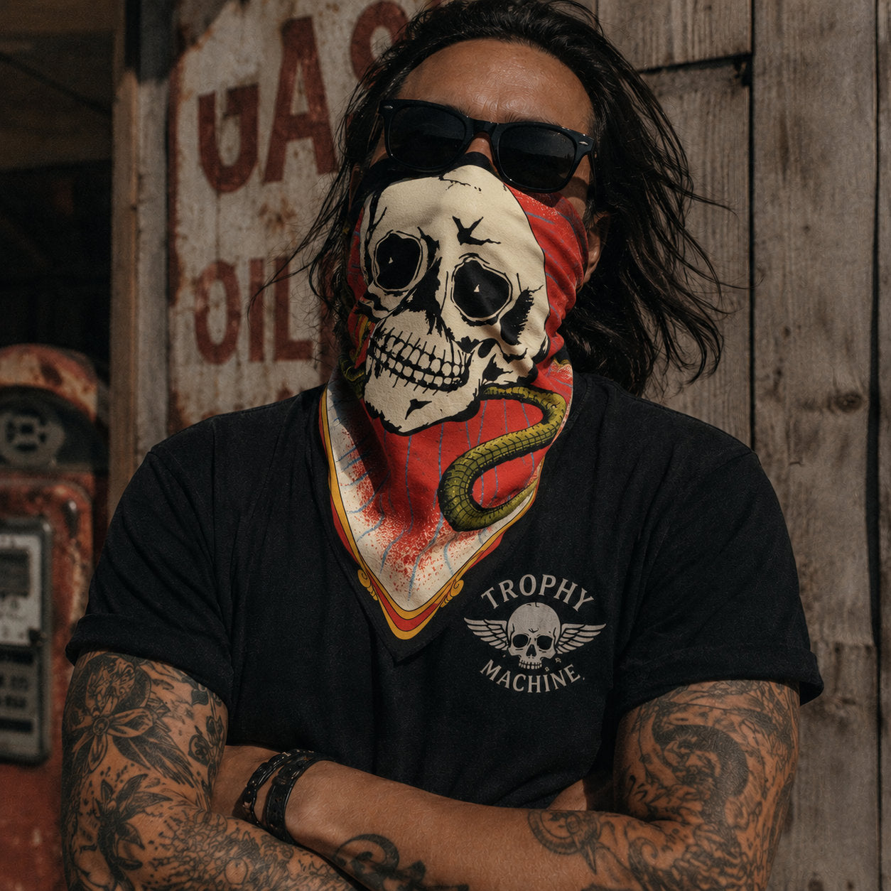 Tattooed man wearing a skull and snake printed bandana folded as a face covering, shot in front of a vintage gas station wall