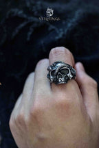 Oxidized silver skull ring with a serpent coiled around the skull, snake head resting on the forehead, worn on a finger against a dark jacket


