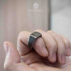 Square signet ring worn on a finger while two hands are clasped together, showing the ring's understated dark silver finish in an intimate everyday context