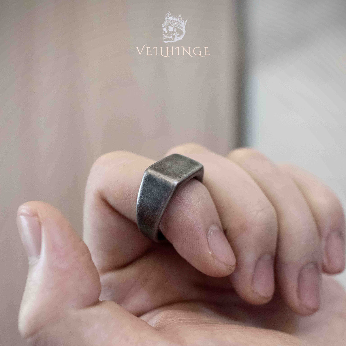 Square signet ring worn on a finger while two hands are clasped together, showing the ring's understated dark silver finish in an intimate everyday context