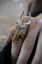 Stag beetle ring with a skull face and blue stone eyes stacked over a gold ribcage band ring, silver beetle with upward horns worn across two fingers against a dark jacket