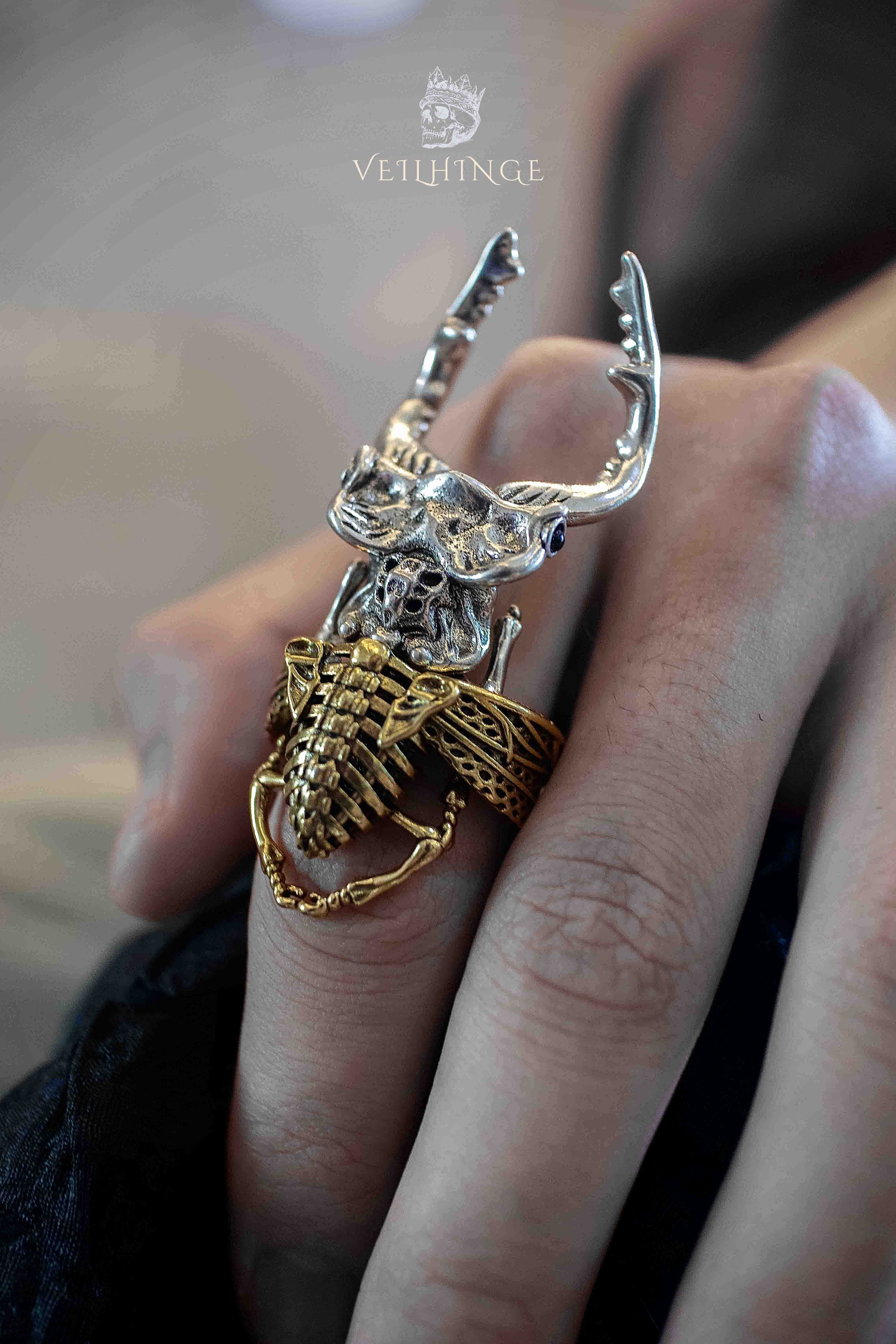 Stag beetle ring with a skull face and blue stone eyes stacked over a gold ribcage band ring, silver beetle with upward horns worn across two fingers against a dark jacket