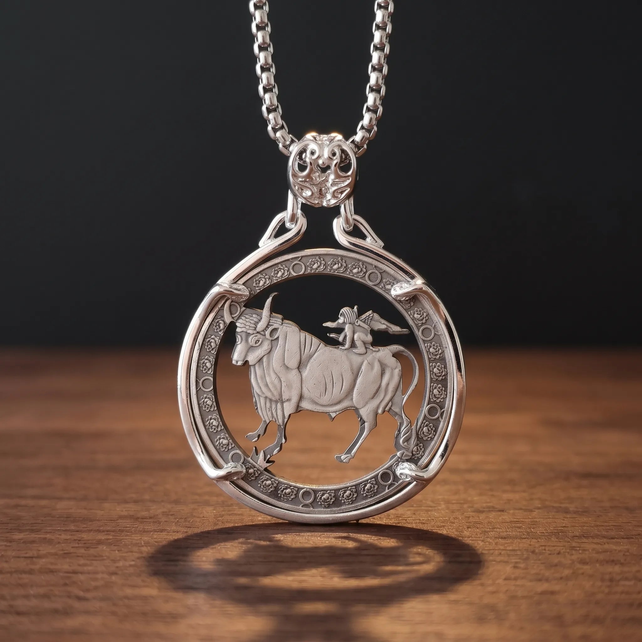 taurus zodiac coin pendant necklace — charging bull with winged cupid figure on back, floral rose border ring, four-prong open frame, ornate skull bail, antique silver tone, warm wood surface