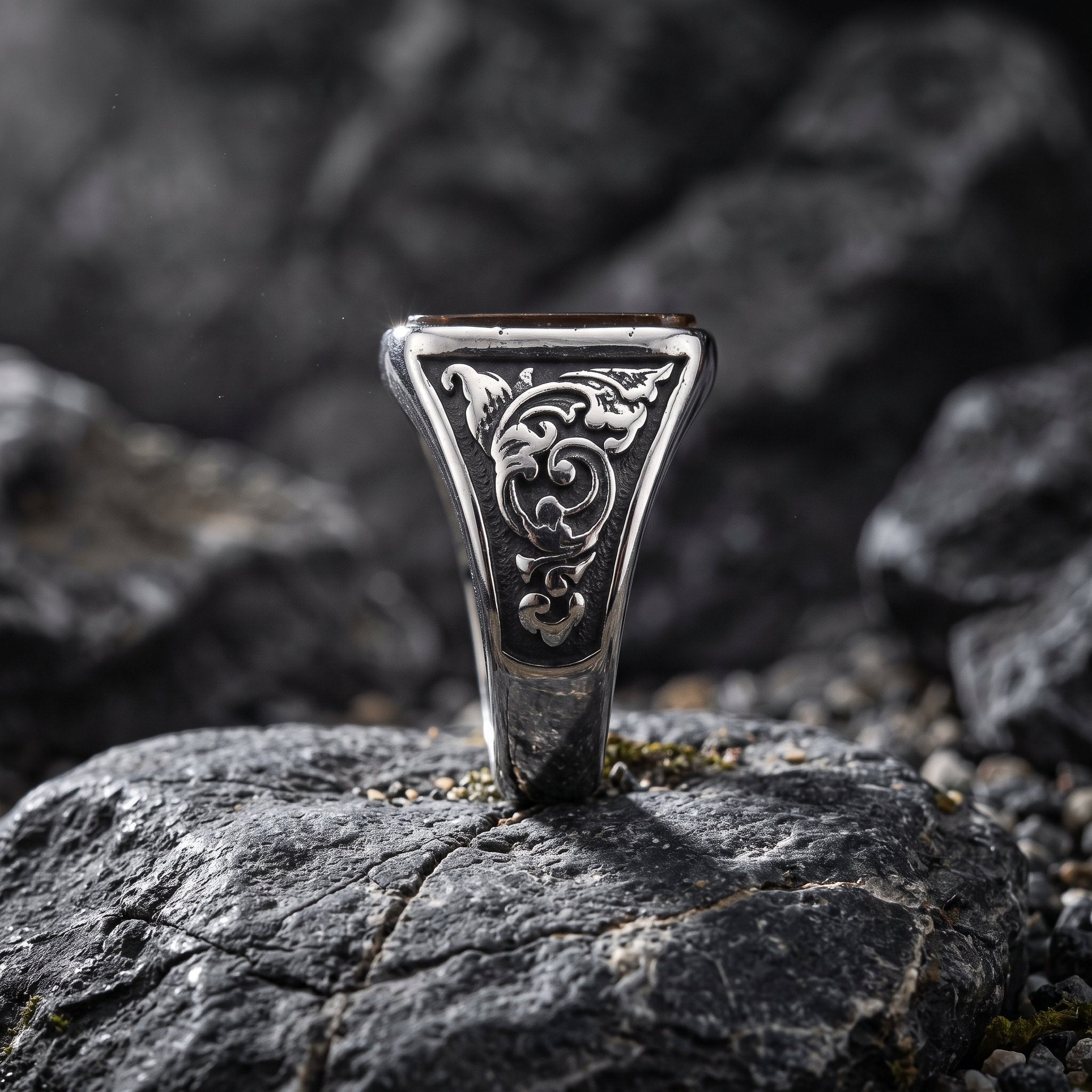 Tiger eye signet ring standing upright in a rock crevice showing the side profile with detailed baroque scroll engraving on the inner band shoulders in dramatic low light