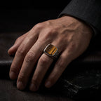 Man's hand wearing a tiger eye signet ring on the ring finger resting on a black leather surface, with the amber and brown stone grain visible in low light