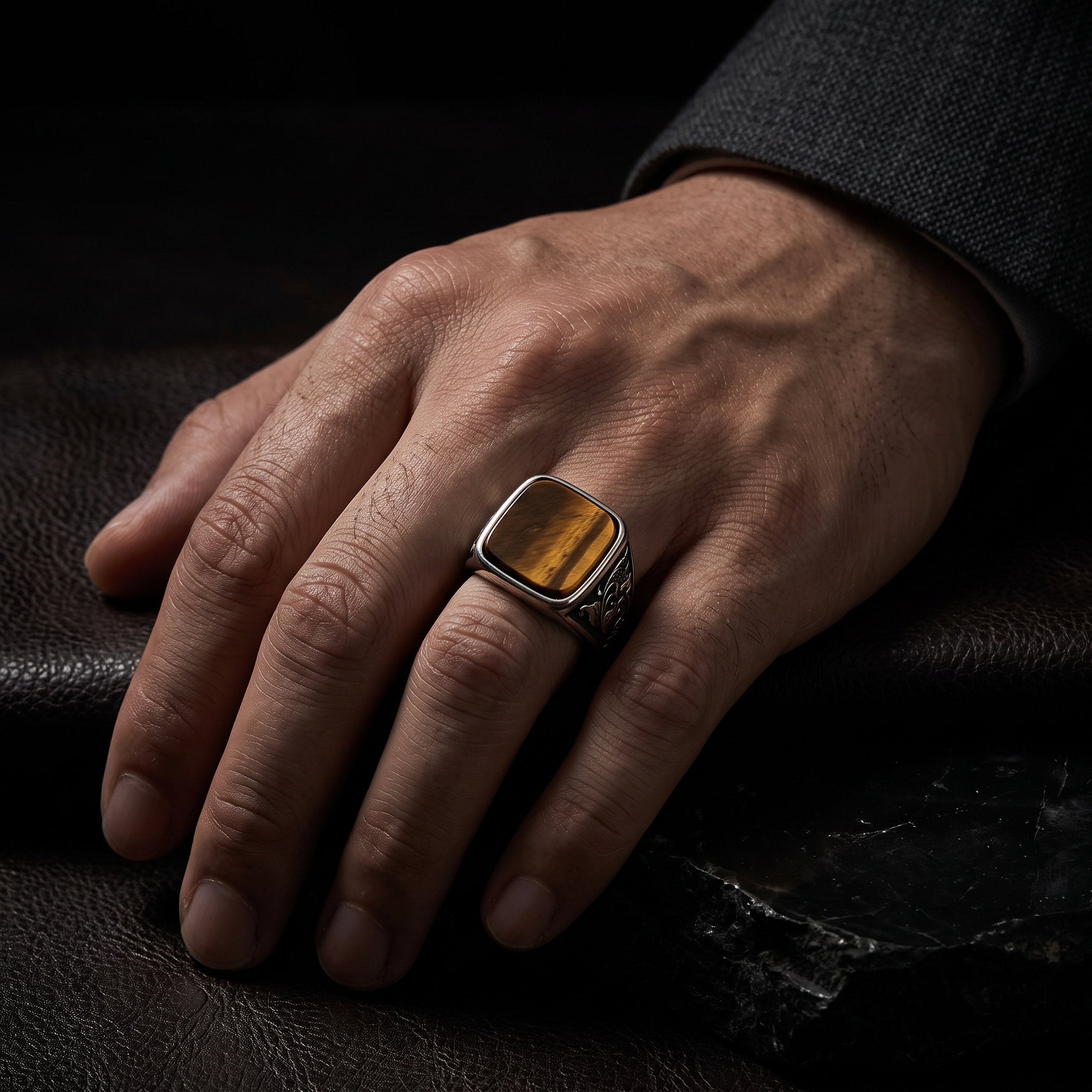 Man's hand wearing a tiger eye signet ring on the ring finger resting on a black leather surface, with the amber and brown stone grain visible in low light