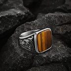 Square tiger eye stone signet ring resting on dark rock, showing the amber brown gemstone face and silver band with baroque floral scroll engraving on both shoulders