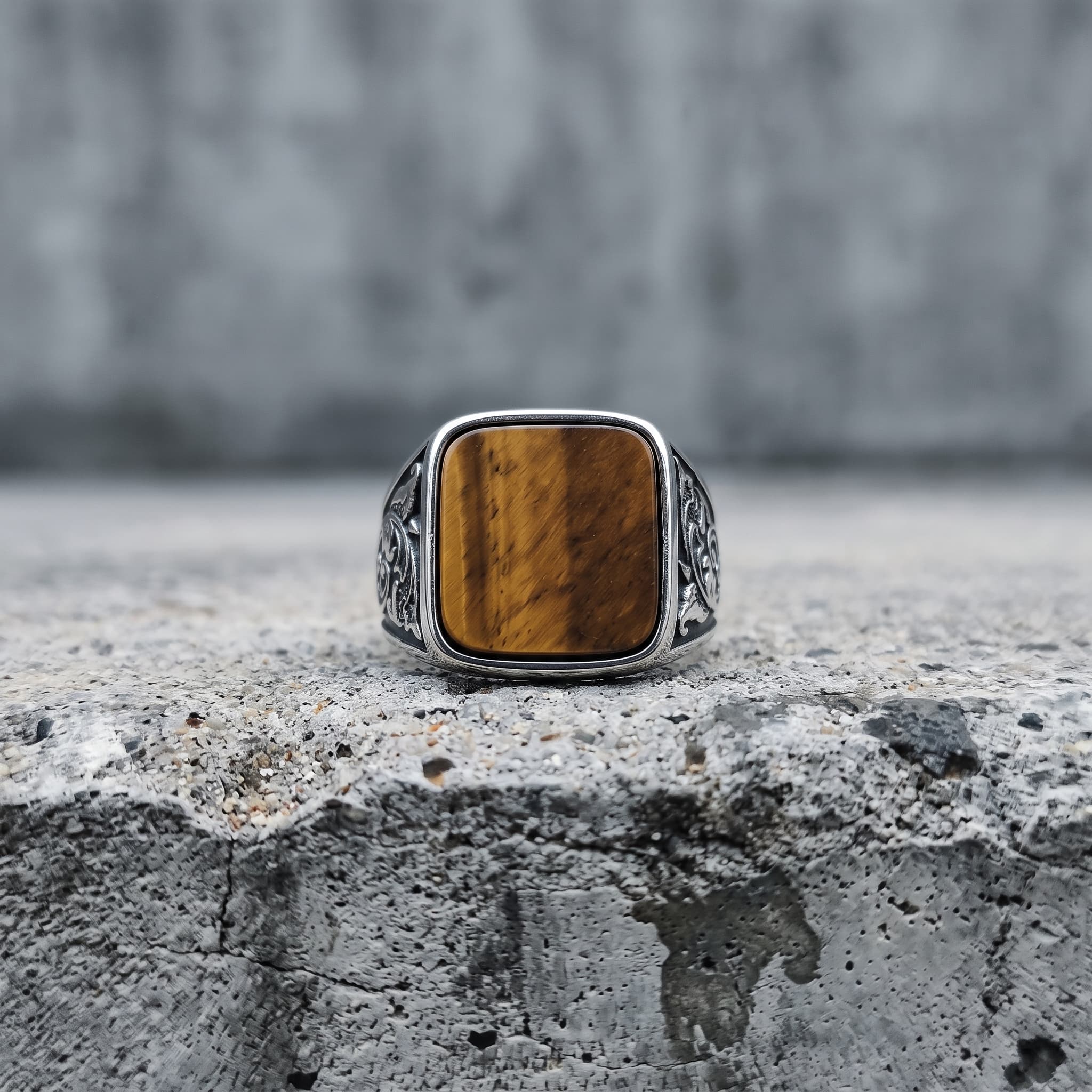 Tiger eye square signet ring standing upright on a concrete ledge with a grey cement wall background, showing the full amber stone face and floral scroll engraving on both band shoulders in natural diffused light Main image