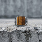 Tiger eye square signet ring standing upright on a concrete ledge with a grey cement wall background, showing the full amber stone face and floral scroll engraving on both band shoulders in natural diffused light