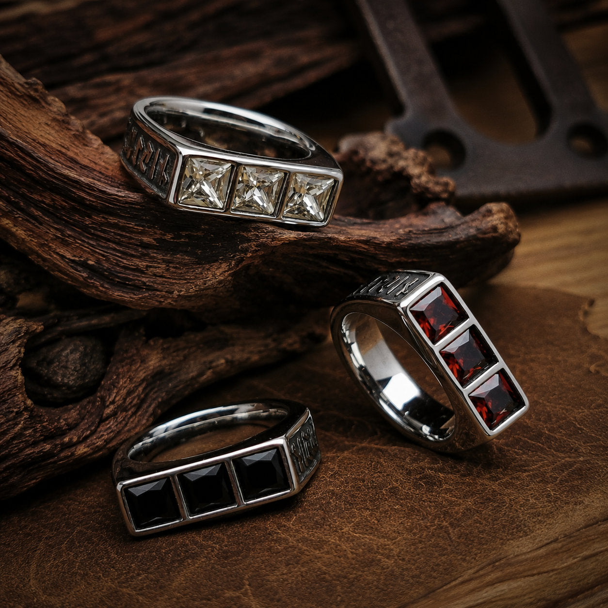 Three square gemstone signet rings arranged on a dark wood and rusted metal surface, showing white crystal, red garnet, and black onyx stone variants with engraved silver bands and dried leaves in the background