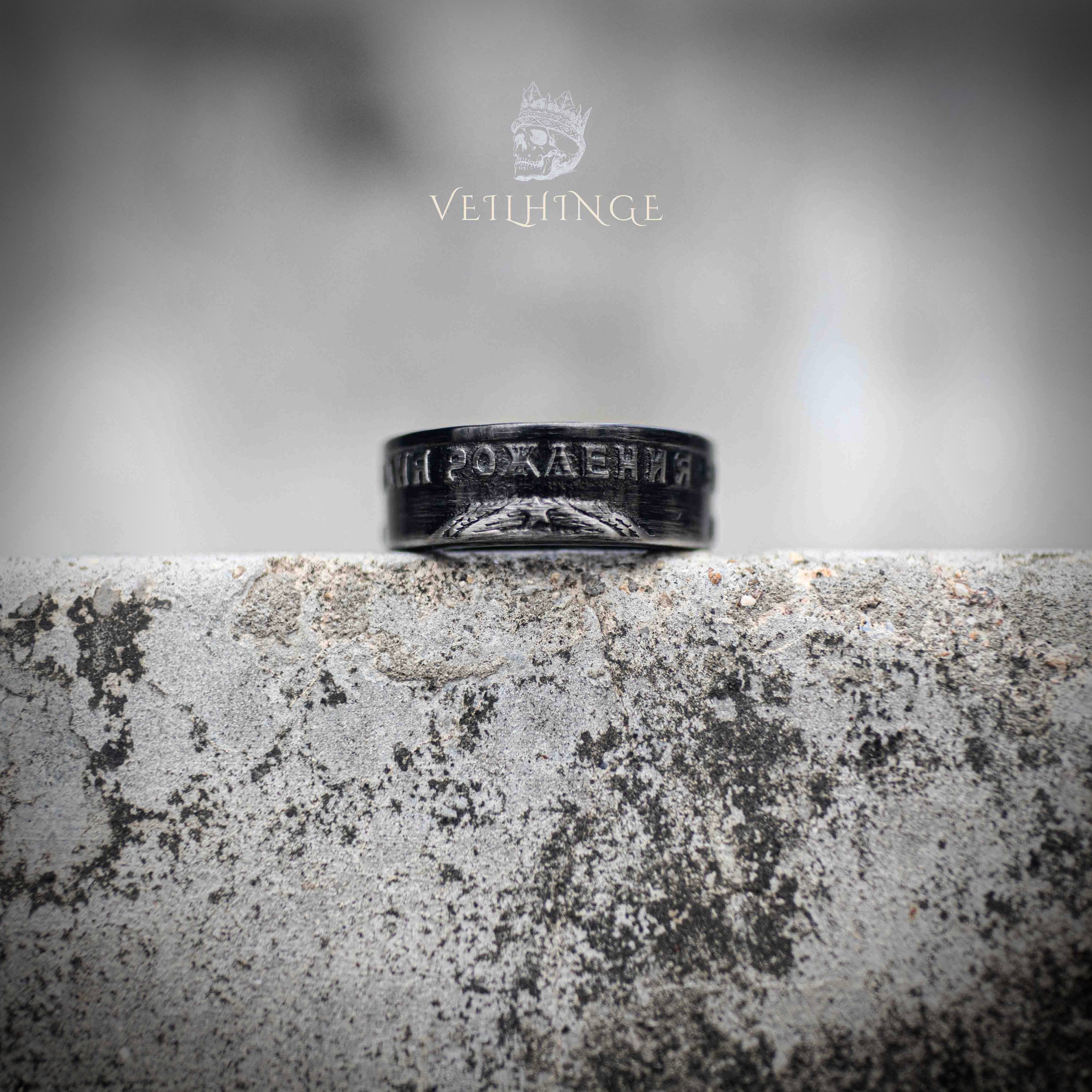 Angled view of black oxidized Soviet coin ring showing partial Cyrillic anniversary text — worn-in dark finish, historical relic aesthetic on weathered stone