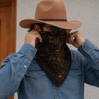 Man pulling a black Wild West bandana up over his face with both hands, wearing a tan cowboy hat and denim western shirt, shot indoors in close-up