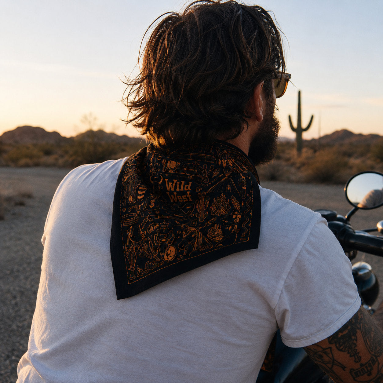 Man riding a motorcycle with a black Wild West bandana draped across the back of his neck, shot from behind against a desert highway and cactus silhouette at sunset