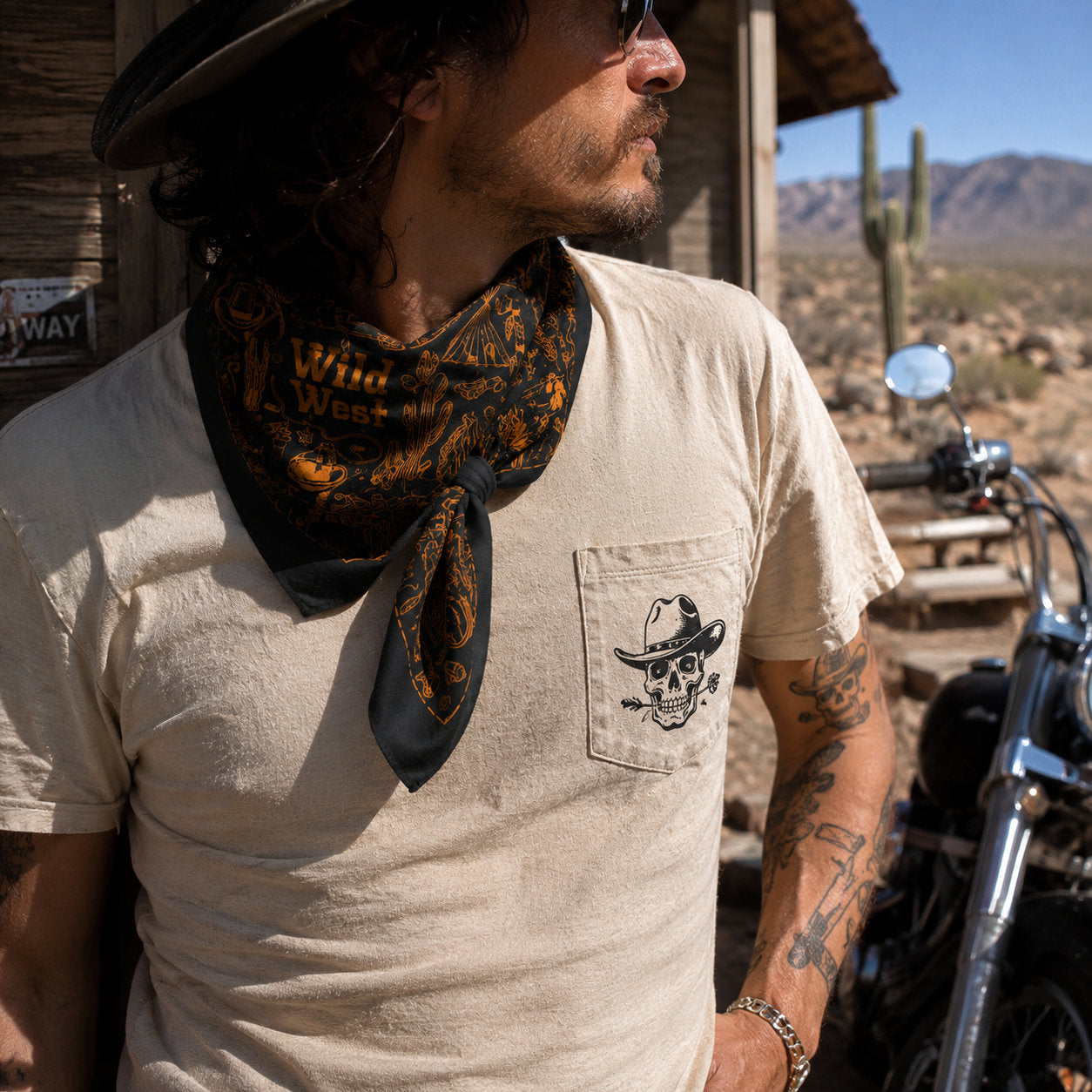 Tattooed man wearing a black and gold Wild West bandana tied around his neck over a white pocket tee, standing beside a motorcycle in a desert setting