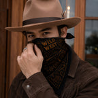 Man wearing a black and gold Wild West bandana as a face covering, styled with a brown fringe leather jacket and cowboy hat, photographed indoors in low light with a ring visible on his hand