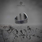 Winged skull signet ring back view on stone surface, Roman numeral band with dense cross engraving, oxidized gunmetal finish, runecore dark nordic jewelry