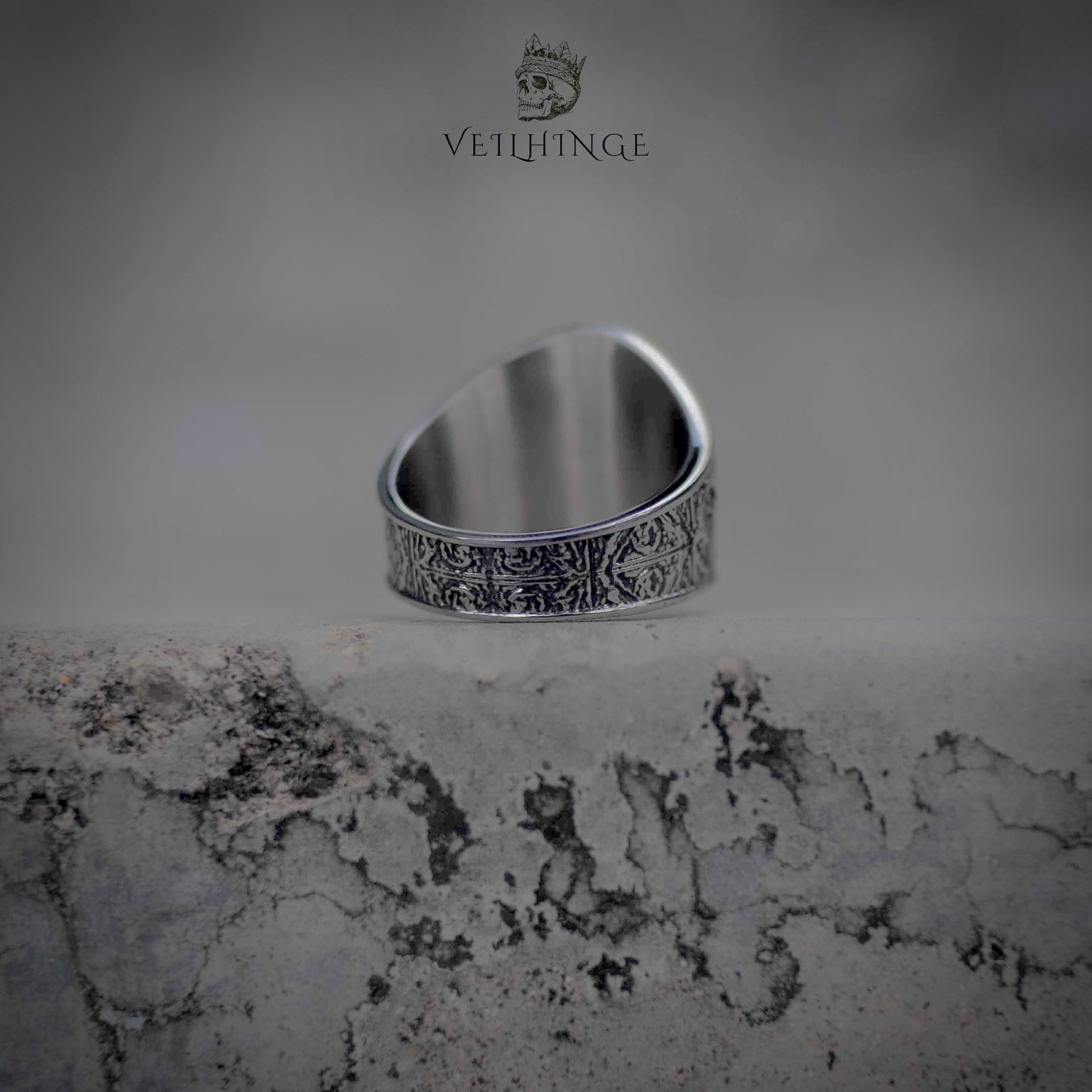 Winged skull signet ring back view on stone surface, Roman numeral band with dense cross engraving, oxidized gunmetal finish, runecore dark nordic jewelry