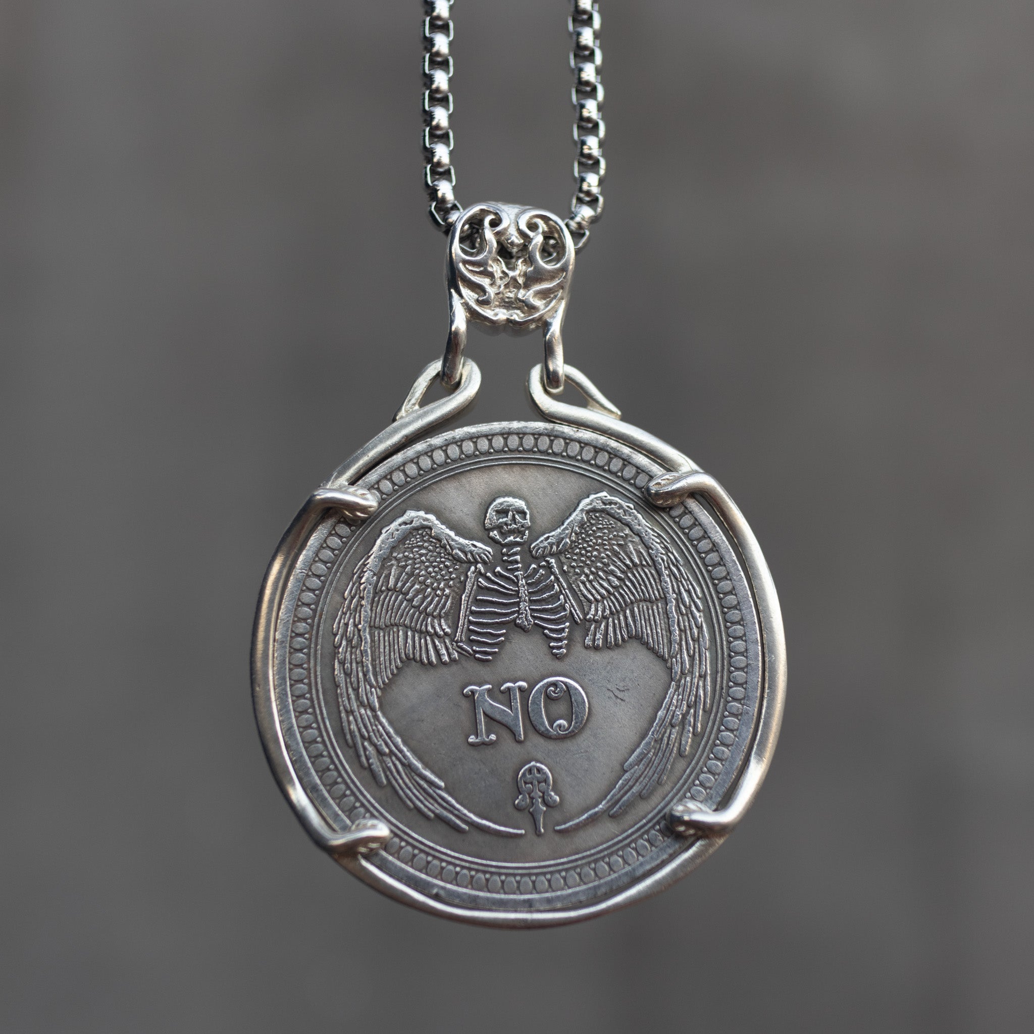 Decision coin pendant showing the NO side with a winged skeleton angel above a heart, set in a four-prong bezel with skull bail on a box chain Main image