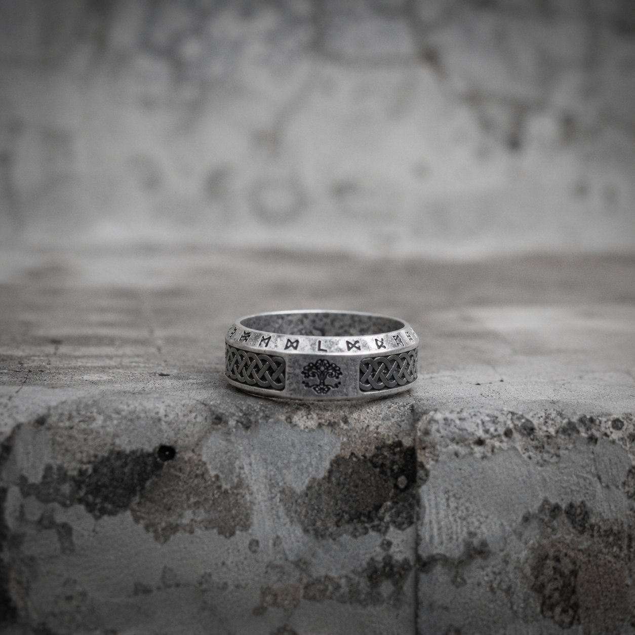 Antique silver Viking ring placed on a concrete ledge, featuring Norse rune engravings and a tree of life motif carved around the band