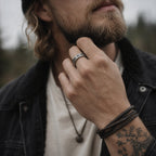 Bearded man wearing an antique silver Yggdrasil ring on his finger, hand raised to his chin, dressed in a black jacket with a leather bracelet and tattoos visible on his wrist

