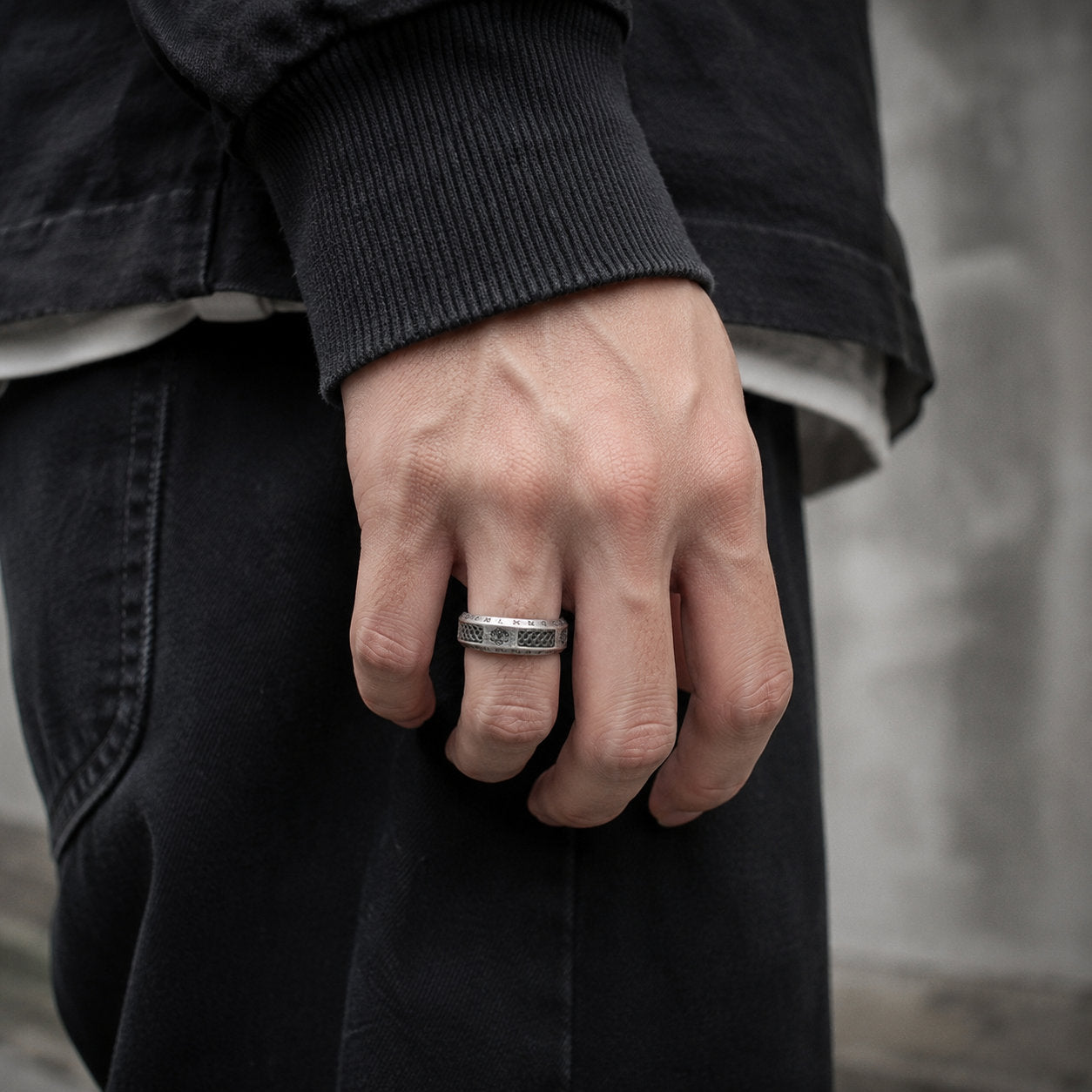 Close-up of a hand wearing an antique silver tree of life Viking ring on the index finger, dressed in a black long-sleeve top with dark trousers in side lighting